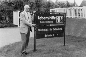 Ein Mann steht vor einem Schild. Er trägt einen Anzug. Das Schild zeigt den Namen eines Ortes.