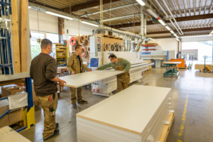 Arbeiter in einer modernen Holzverarbeitungswerkstatt, die große Platten bearbeiten und vorbereiten. Die Umgebung zeigt Maschinen und Werkzeuge für die Holzverarbeitung.