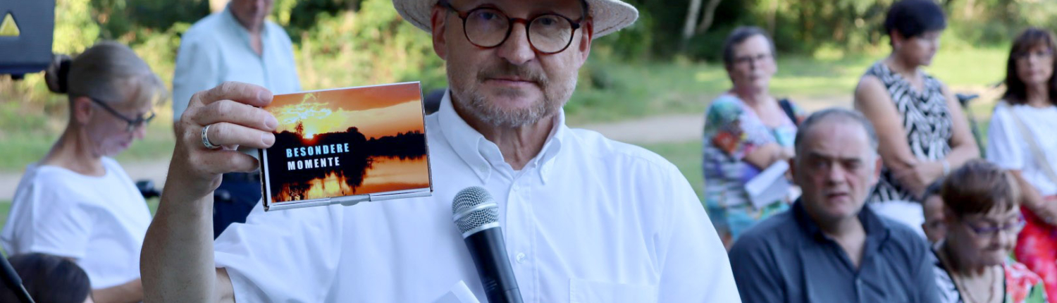 Mann mit Hut hält ein Dokument in der Hand und spricht in ein Mikrofon, während eine Gruppe von Zuhörern im Hintergrund sitzt, umgeben von Bäumen.