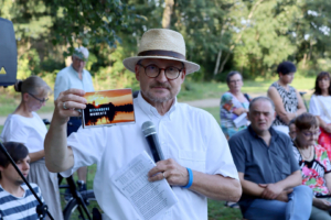 Mann mit Hut hält ein Dokument in der Hand und spricht in ein Mikrofon, während eine Gruppe von Zuhörern im Hintergrund sitzt, umgeben von Bäumen.