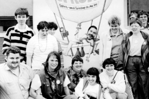 Gruppe von Menschen in Schwarz-Weiß-Foto, die vor einem Banner stehen, das eine Veranstaltung oder Kampagne unterstützt. Einige Personen tragen T-Shirts mit Symbolen oder Slogans. Die Atmosphäre wirkt freundlich und engagiert.