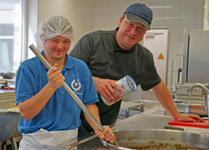 Zwei Mitarbeiter in einer Lebensmittelverarbeitungseinrichtung, die gemeinsam an einem großen Kochtopf arbeiten. Einer der Mitarbeiter hält einen Löffel, während der andere eine Zutat hinzufügt. Beide tragen Schutzkleidung, einschließlich Haarnetz und Schürze.