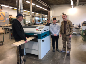 Vier Personen stehen in einer Werkstatt neben einer modernen Holzverarbeitungsmaschine. Sie halten ein Holzbrett und zeigen Interesse an der Maschine. Die Umgebung ist gut beleuchtet und zeigt eine professionelle Arbeitsatmosphäre.