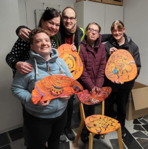 Gruppe von fünf Personen, die stolz bemalte Stühle mit bunten, kreativen Designs präsentieren, in einem hellen Raum mit grauem Boden.