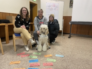 Drei Frauen sitzen mit einem Hund in einem Seminarraum. Vor ihnen liegen bunte Buchstabenkarten. Der Hund, ein Pudel, sitzt aufmerksam in der Mitte.
