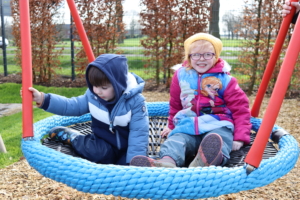 Zwei Kinder sitzen auf einer Schaukel. Ein Kind hat eine blaue Jacke an, das andere trägt rot. Sie sitzen draußen auf einem Spielplatz.