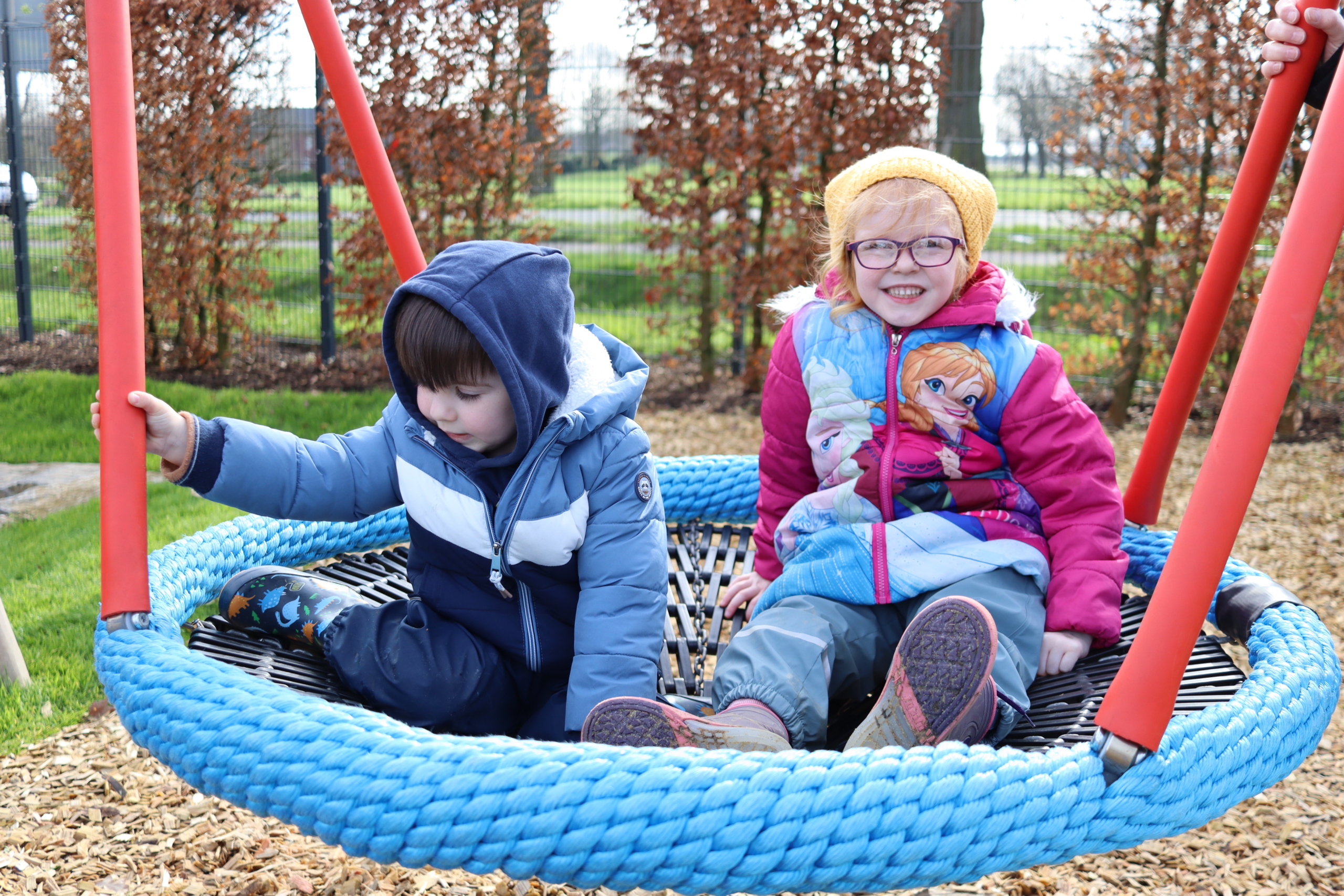 Zwei Kinder sitzen auf einer Schaukel. Ein Kind hat eine blaue Jacke an, das andere trägt rot. Sie sitzen draußen auf einem Spielplatz.