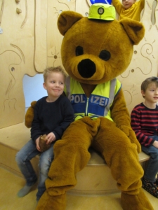 Ein großer, freundlicher Bär in Polizeikleidung sitzt zwischen zwei lächelnden Kindern in einem spielerischen Umfeld.
