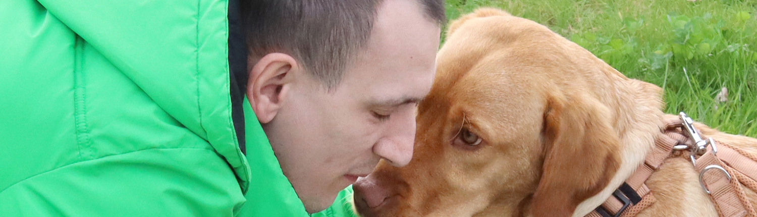 Mann in grüner Jacke, der sich liebevoll mit einem Labrador-Hund beschäftigt, während sie auf einer Wiese spielen.