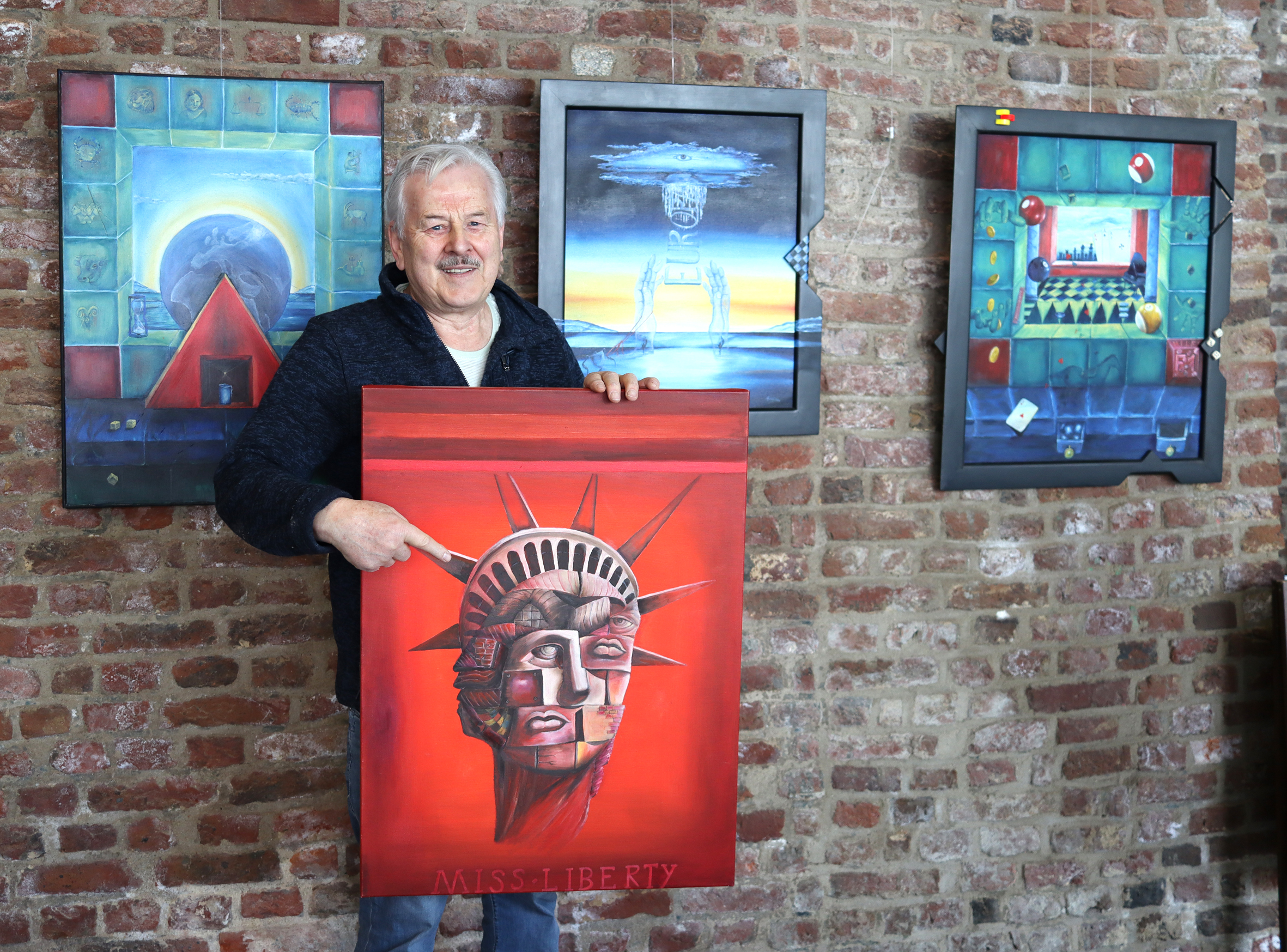 Ein Künstler hält ein rot gefärbtes Gemälde mit einem Gesicht im Zentrum, umgeben von mehreren Kunstwerken an einer Backsteinwand.