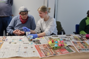 Zwei Kinder bemalen Eier mit Farbe. Sie sitzen an einem Tisch mit Papier.