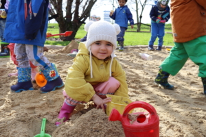 Kleines Mädchen im gelben Regenmantel und weißen Mütze, das im Sandkasten spielt und mit einem roten Gießkanne spielt, umgeben von anderen Kindern und Spielzeug im Freien.