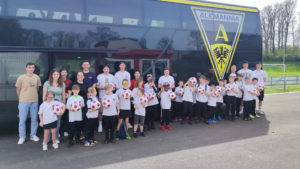 Gruppe von Kindern und Trainern in weißen T-Shirts mit Farben des Vereins, versammelt vor einem Vereinsbus, während einer Sportveranstaltung oder Trainingseinheit.