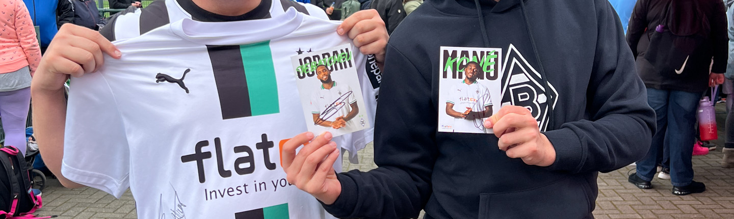 Zwei Männer stehen draußen. Einer trägt ein Fußballtrikot und der andere eine schwarze Jacke. Sie halten etwas in der Hand.