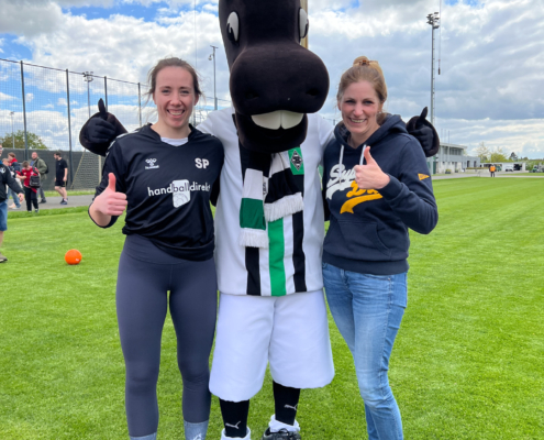 Zwei Frauen stehen auf einem Sportplatz. Sie tragen Sportklamotten und lächeln. Im Hintergrund sind noch andere Menschen.