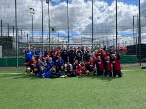 Gruppenfoto von zwei Fußballmannschaften in verschiedenen Trikots auf einem Sportplatz, umgeben von Toren und einem blauen Himmel mit Wolken.