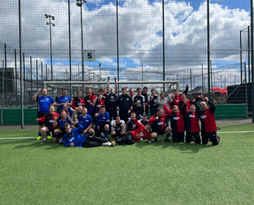Gruppenfoto von zwei Fußballmannschaften in verschiedenen Trikots auf einem Sportplatz, umgeben von Toren und einem blauen Himmel mit Wolken.