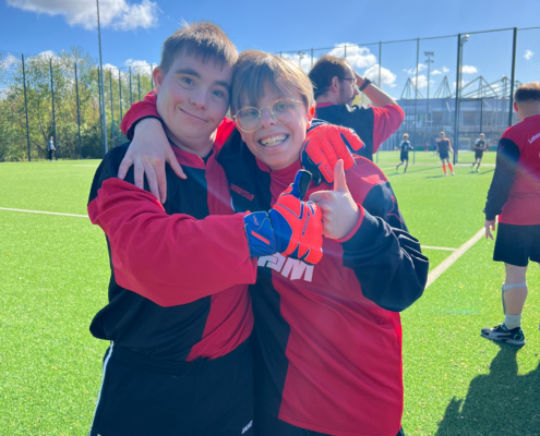 Zwei Jungen stehen auf einem Fußballplatz. Einer hat ein rotes Trikot an, der andere ein schwarzes Trikot. Beide lächeln.