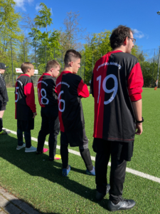 Fünf Fußballspieler in schwarz-roten Trikots stehen auf einem Sportfeld und warten auf den Spielbeginn. Die Rückennummern 6, 8, 9 und 19 sind sichtbar.