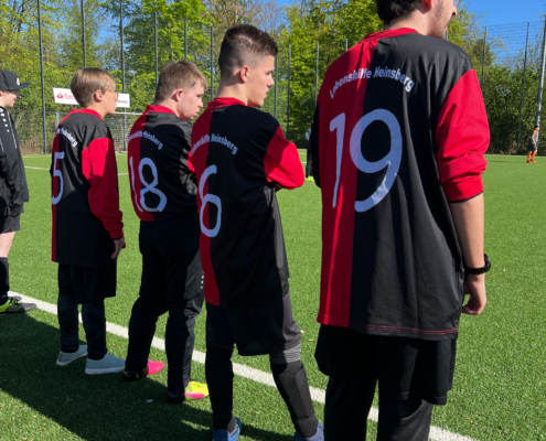 Fünf Fußballspieler in schwarz-roten Trikots stehen auf einem Sportfeld und warten auf den Spielbeginn. Die Rückennummern 6, 8, 9 und 19 sind sichtbar.