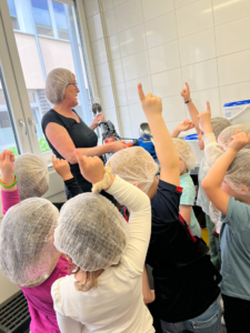 Lehrerin erklärt Kindern im Vorschulalter ein Thema, während die Kinder mit erhobenen Händen Fragen stellen. Die Kinder tragen Haarnetze und sind in einem hellen Klassenraum.