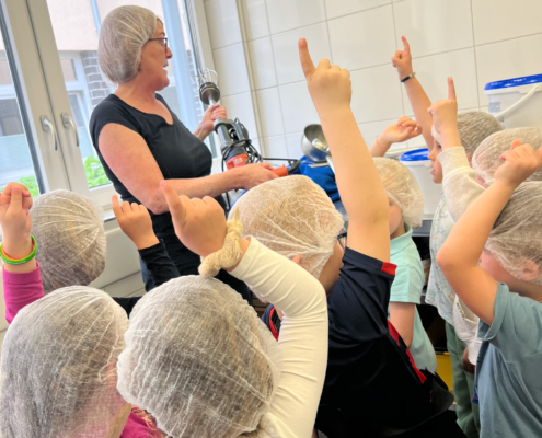2024-05-16_Besuch Küche 1 (28)-2 Lehrerin erklärt Kindern im Vorschulalter ein Thema, während die Kinder mit erhobenen Händen Fragen stellen. Die Kinder tragen Haarnetze und sind in einem hellen Klassenraum.