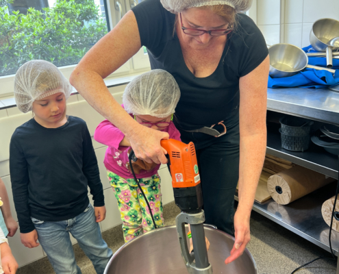 2024-05-16_Besuch Küche 1 (35)-2 Eine Frau rührt mit einem großen Mixer etwas in einem Behälter. Zwei Kinder stehen daneben.