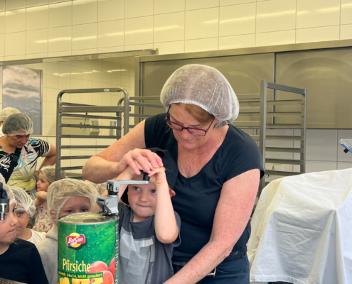 2024-05-16_Besuch Küche 1 (41)-2 Eine Frau mit Haarnetz und einem Kind in einer Küche, die gemeinsam an einer großen Dose arbeiten. Im Hintergrund sind Backbleche und Küchenutensilien sichtbar.