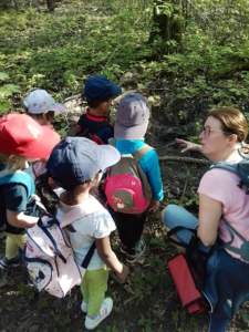 Kinder stehen im Wald. Eine Frau sitzt auf dem Boden und spricht mit ihnen.