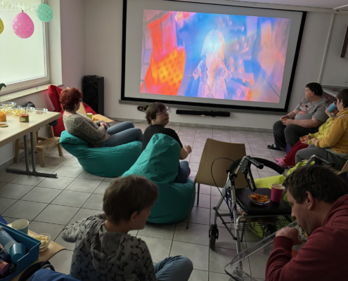 6 Die Bewohner der Wohnstätte machen Kino in ihrem Zuhause. Eine Leinwand ist aufgebaut und sie sitzen davor auf dem Boden.