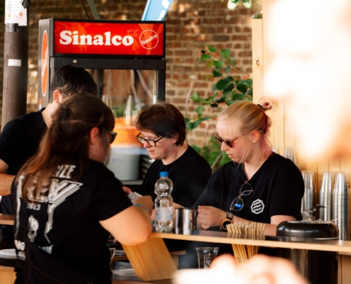 Drei Personen stehen hinter einer Bar. Sie tragen schwarze T-Shirts und eine trägt eine Brille.