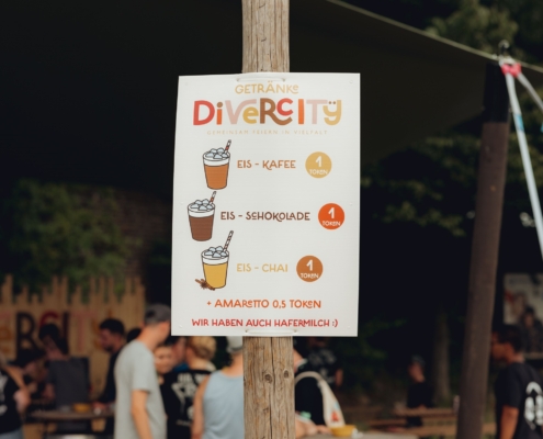 Ein Mann hält ein Getränk. Im Hintergrund sind Menschen. Es ist draußen.