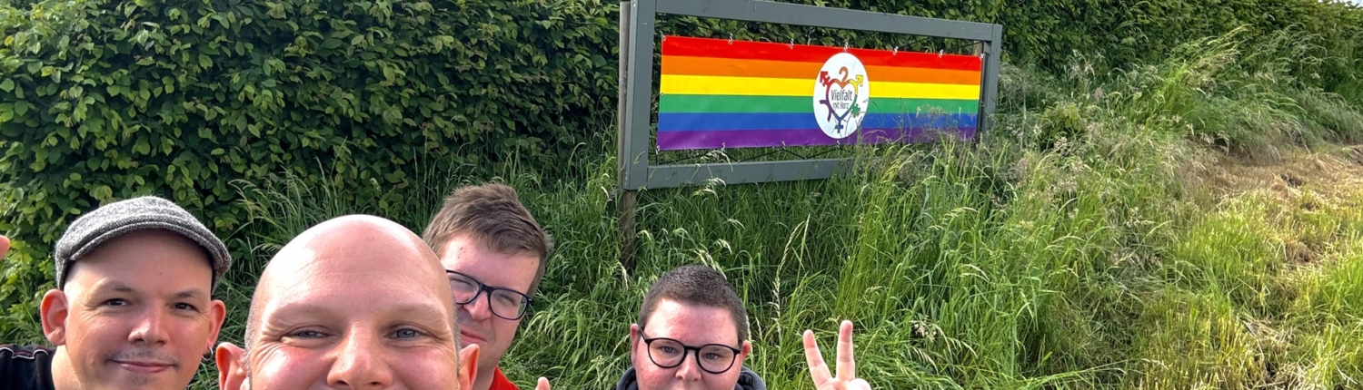 Gruppe von vier Personen posiert fröhlich vor einem Schild mit einer Regenbogenflagge, das Vielfalt und Inklusion symbolisiert. Im Hintergrund ist eine grüne Landschaft zu sehen.