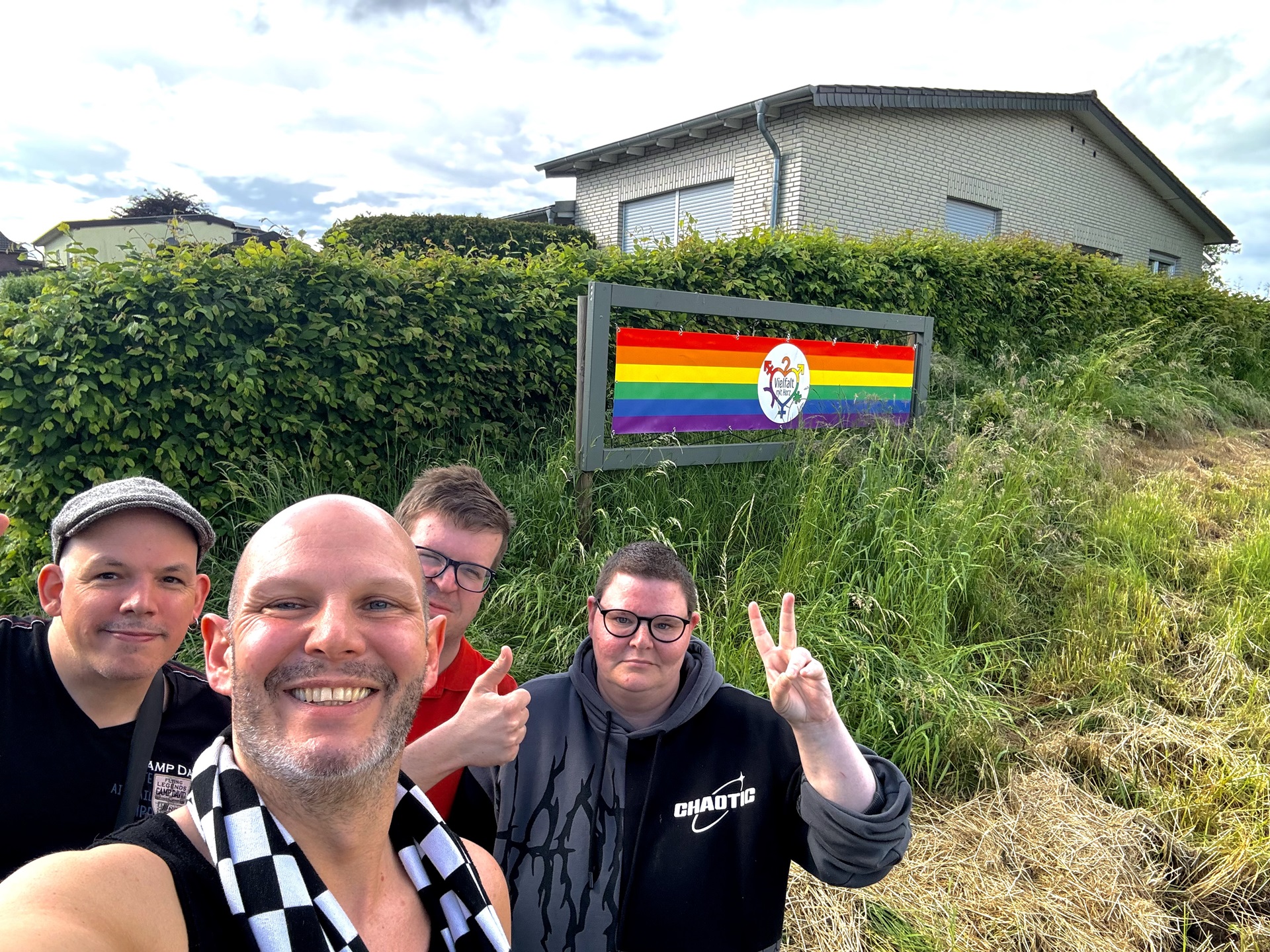 Gruppe von vier Personen posiert fröhlich vor einem Schild mit einer Regenbogenflagge, das Vielfalt und Inklusion symbolisiert. Im Hintergrund ist eine grüne Landschaft zu sehen.