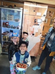 Eine Gruppe von drei Kindern und einer Erwachsenen posiert lächelnd vor einem Kühlschrank und einer Holzregalwand. Die Kinder tragen lässige Kleidung und die Erwachsene hat eine Jeansjacke an. Die Szene strahlt Freude und Zusammengehörigkeit aus.