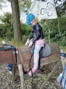 Ein Kind in einem blauen Hut sitzt auf einem Holzstück und übt das Reiten. Im Hintergrund sind Bäume und eine beschilderte Fläche zu sehen, die eine ländliche Umgebung zeigt.