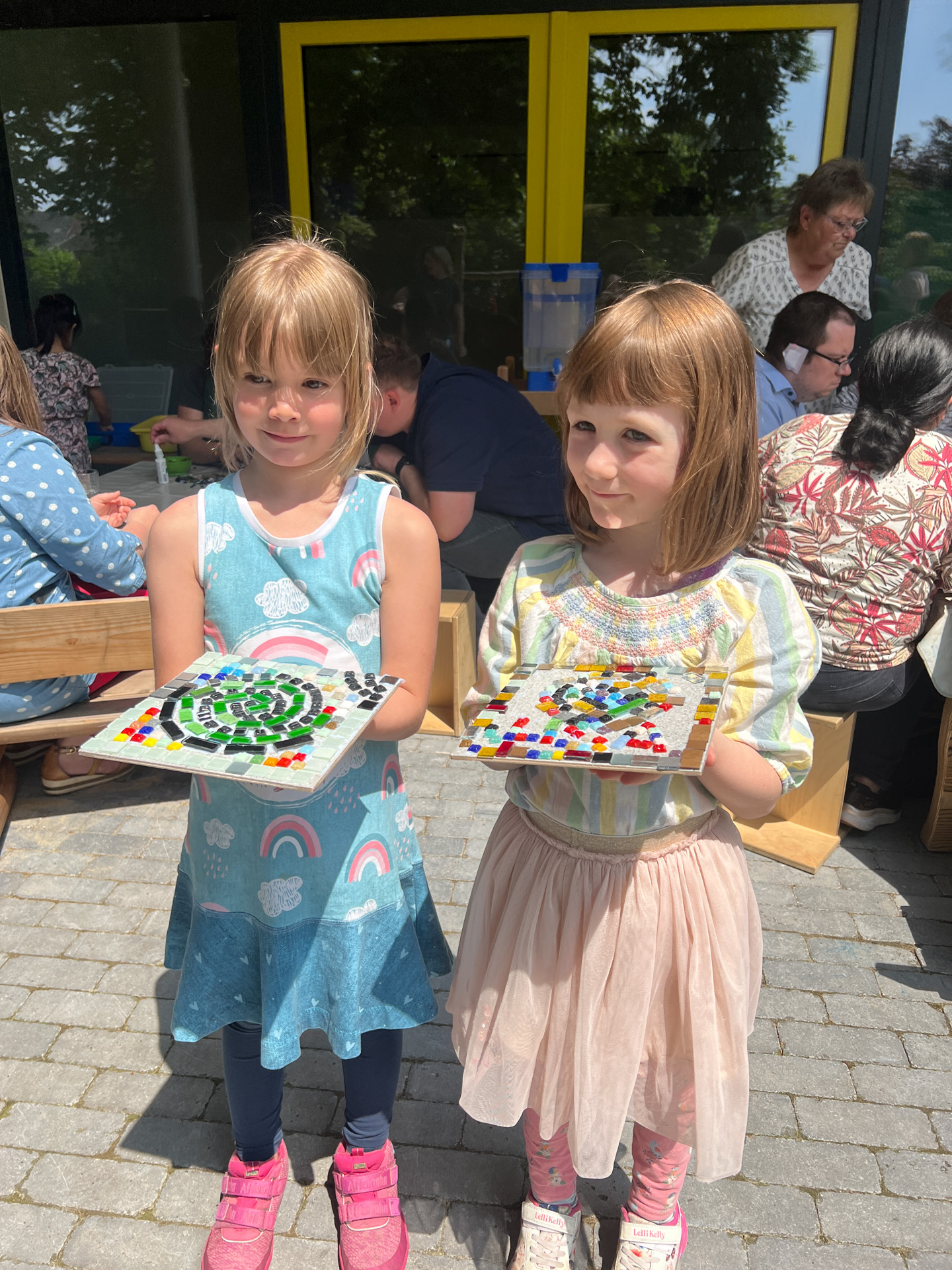 Zwei Mädchen halten Bilder mit bunten Steinen. Sie lächeln.