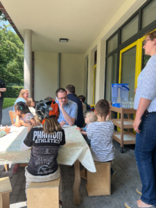 Kinder sitzen an einem Tisch im Freien. Ein Mann steht daneben und spricht mit ihnen.