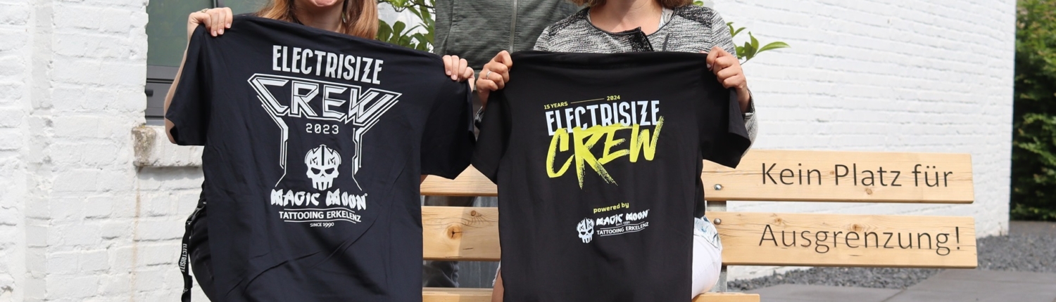 Drei Personen, die auf einer Bank sitzen und T-Shirts mit verschiedenen Aufdrucken tragen, posieren fröhlich vor einer modernen Gebäudeoberfläche. Die Szene vermittelt ein Gefühl von Gemeinschaft und Freizeit.