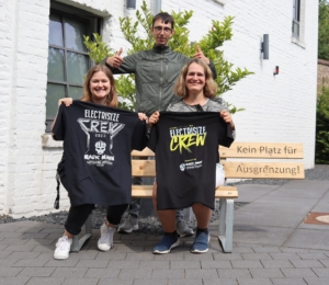Drei Personen, die auf einer Bank sitzen und T-Shirts mit verschiedenen Aufdrucken tragen, posieren fröhlich vor einer modernen Gebäudeoberfläche. Die Szene vermittelt ein Gefühl von Gemeinschaft und Freizeit.