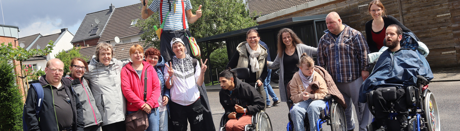 Eine Gruppe Menschen steht vor einem Gebäude. Ein Mann mit Clown-Kostüm hält einen Regenschirm. Einige sitzen im Rollstuhl.