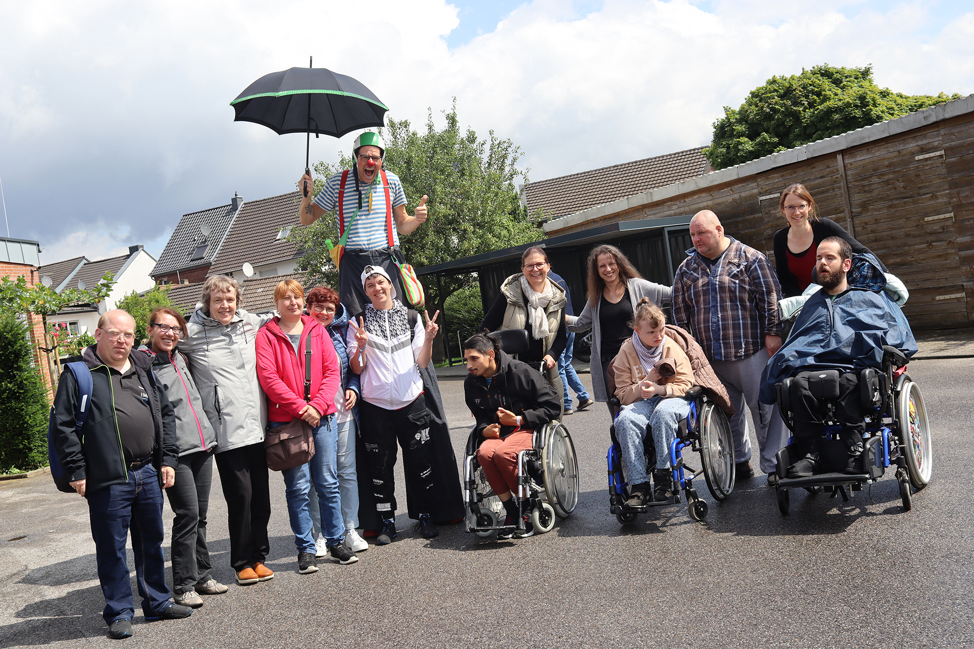 Eine Gruppe Menschen steht vor einem Gebäude. Ein Mann mit Clown-Kostüm hält einen Regenschirm. Einige sitzen im Rollstuhl.