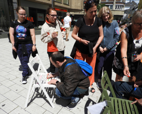 2024-07-14_Jeder braucht einen Platz im Leben Party (16)-2 Ein Mann kniet auf dem Gehweg und schreibt etwas. Mehrere Leute schauen zu.