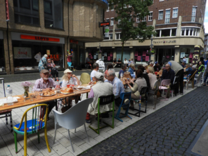 Viele Leute sitzen an einem langen Tisch draußen und essen. Es sieht nach einer Party oder einem Fest aus.