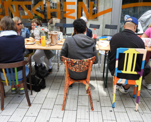 2024-07-14_Jeder braucht einen Platz im Leben Party (38)-2 Personen sitzen an einem Tisch im Freien, umgeben von bunten Stühlen und einer modernen Café-Atmosphäre. Im Hintergrund ist ein kreatives Schriftzug sichtbar.