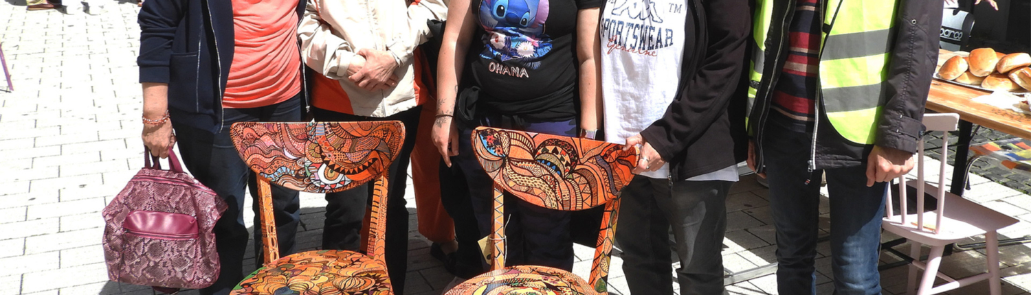 Gruppe von Menschen steht lächelnd vor zwei dekorierten Stühlen auf einem Platz. Die Szene zeigt eine freundliche Atmosphäre und Gemeinschaftsgeist.