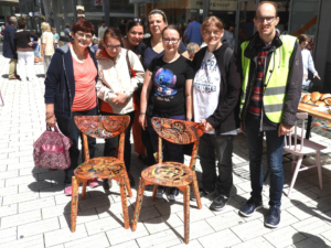 Gruppe von Menschen steht lächelnd vor zwei dekorierten Stühlen auf einem Platz. Die Szene zeigt eine freundliche Atmosphäre und Gemeinschaftsgeist.