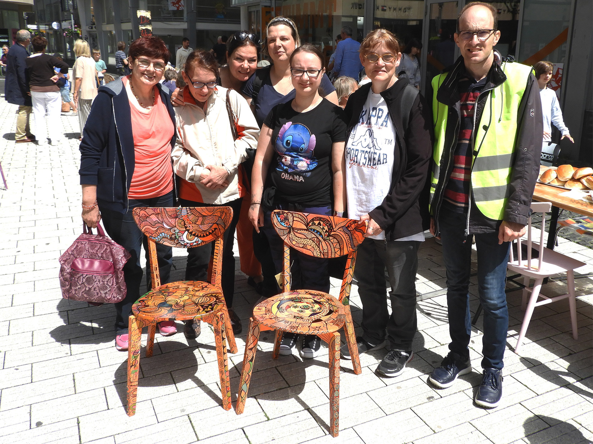 Gruppe von Menschen steht lächelnd vor zwei dekorierten Stühlen auf einem Platz. Die Szene zeigt eine freundliche Atmosphäre und Gemeinschaftsgeist.