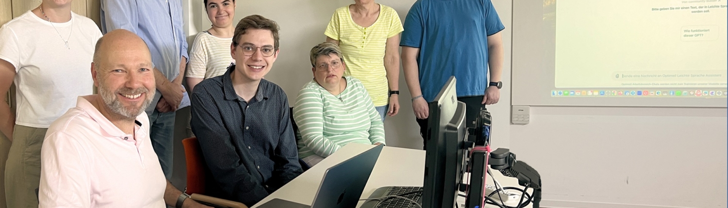 Gruppe von sechs Personen in einem modernen Büroraum, die an einem Tisch mit Laptops und technischen Geräten zusammenarbeiten. Helle, freundliche Atmosphäre mit großen Fenstern und einem Bildschirm im Hintergrund.