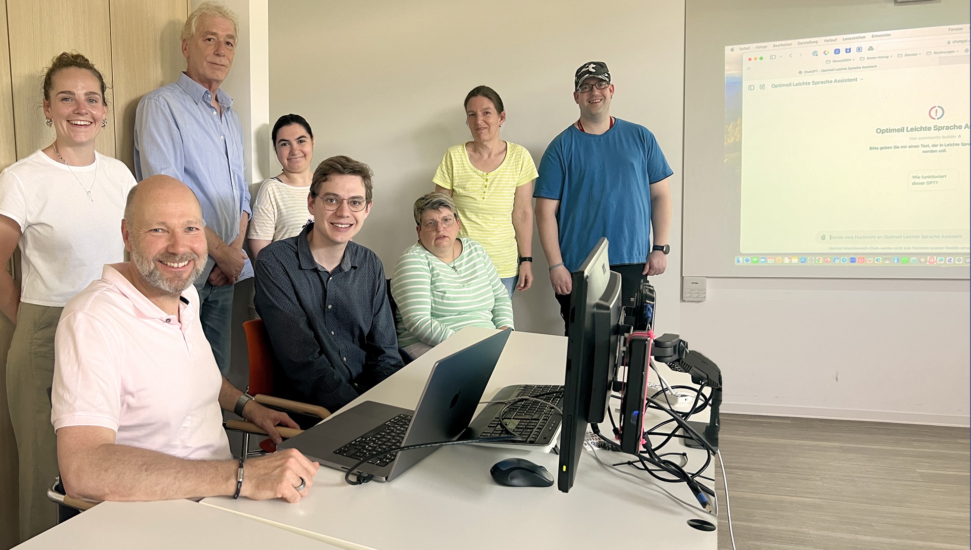 Gruppe von sechs Personen in einem modernen Büroraum, die an einem Tisch mit Laptops und technischen Geräten zusammenarbeiten. Helle, freundliche Atmosphäre mit großen Fenstern und einem Bildschirm im Hintergrund.